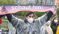 Ein junger Mann mit einem Regenponcho hebt die Arme und hält den Kirchentagsschal hoch