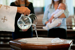 Hand gießt Wasser ins Taufbecken, im Hintergrund Erwachsene mit Baby am Arm