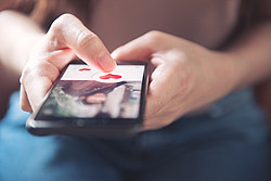 Hand einer Frau hält Handy in der Hand, auf dem ist das Bild es eines Mannes zu erahnen und ein rotes Herz zu sehen