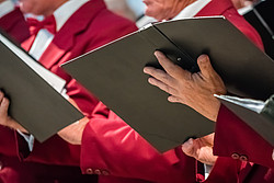 Zwei Menschen singen und halten Notenbücher in der Hand. Sie tragen weiße Hemden, rote Fliegen und rote Jacketts.Ihre Gesichter sind nicht zu sehen.