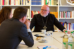 Kardinal Marx sitzt am Tisch und spricht mit einer Frau und einem Mann, von denen nur der Rücken zu sehen ist.